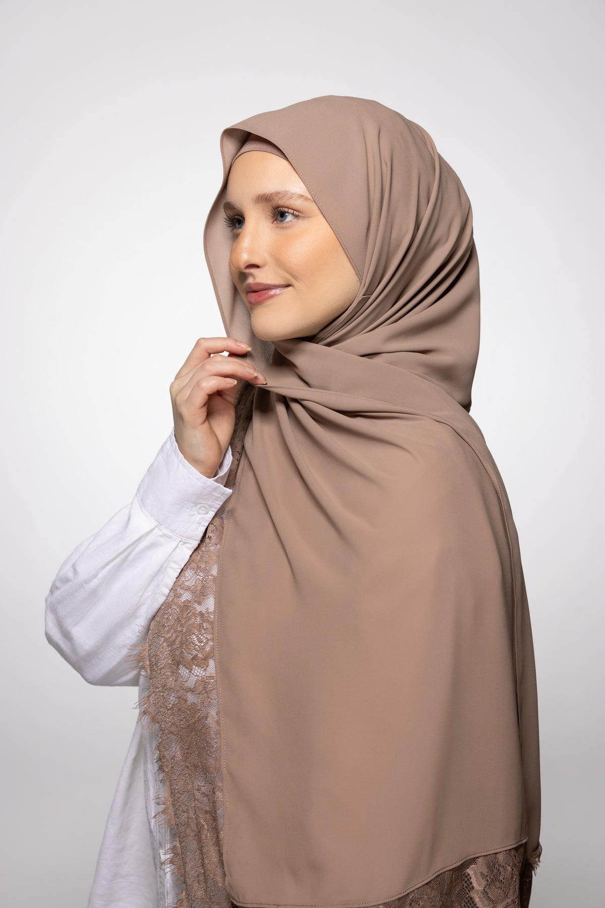 A woman wearing a draped taupe-brown hijab and a white lace-trimmed top, photographed against a clean light gray studio background.