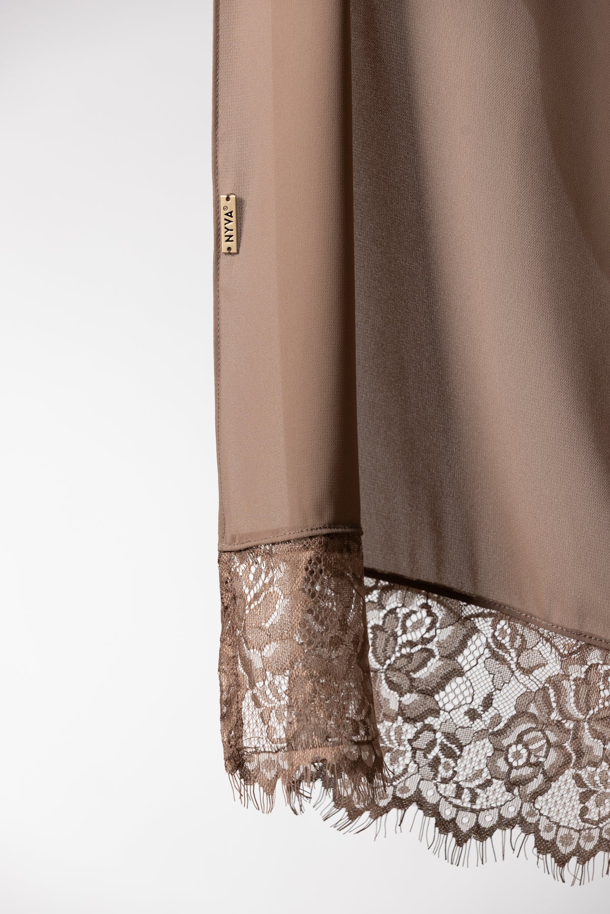 Close-up of a taupe-brown long-sleeve garment with a sheer floral lace hem and a small gold-toned tag reading NYVA.