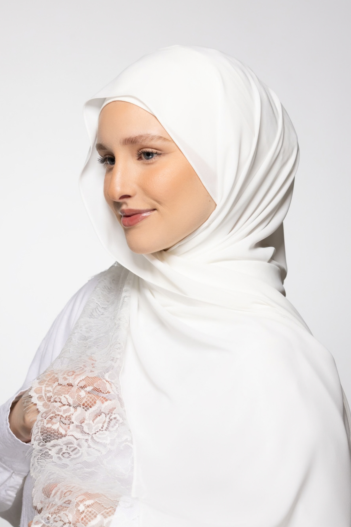A woman wearing a white hijab that is lace-trimmed, photographed against a clean light gray studio background.