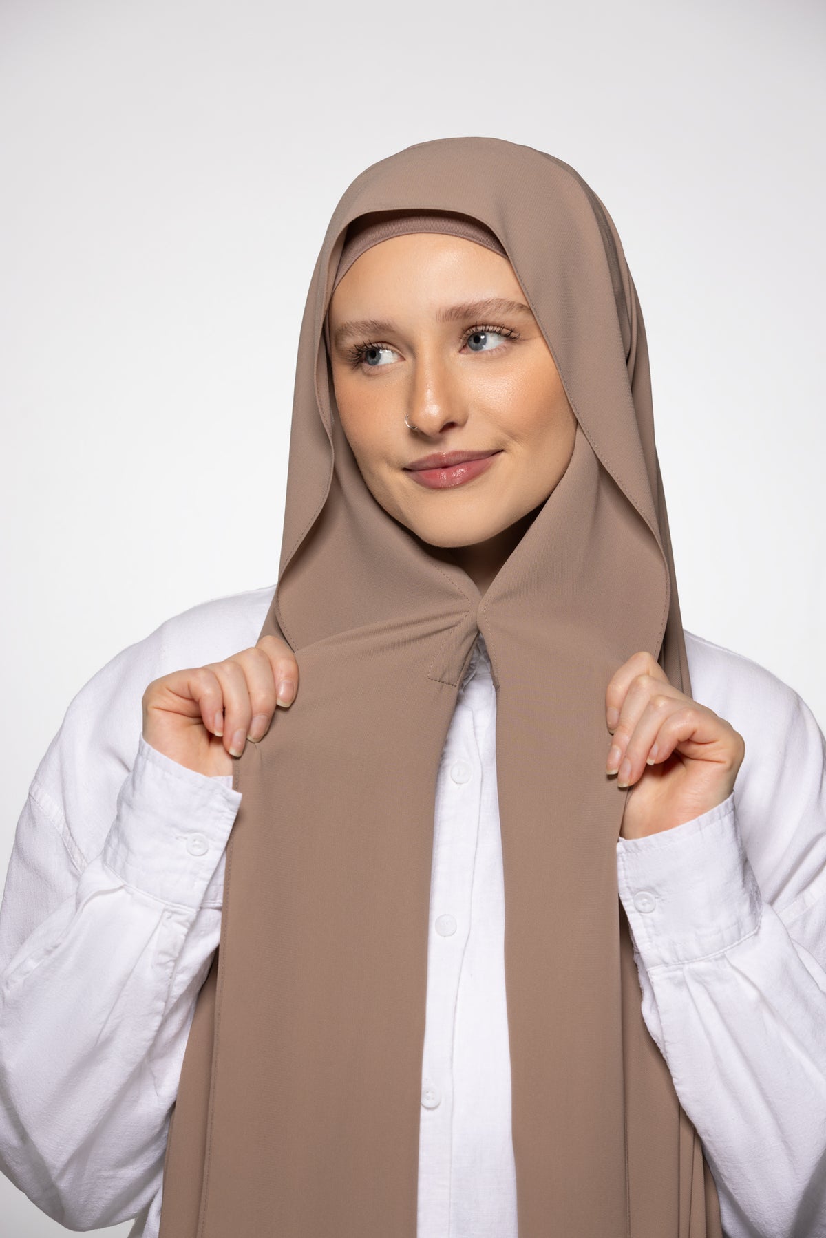 A woman wearing a taupe brown hijab and a white button-up shirt, holding the front of the hijab with both hands against a light gray studio background.