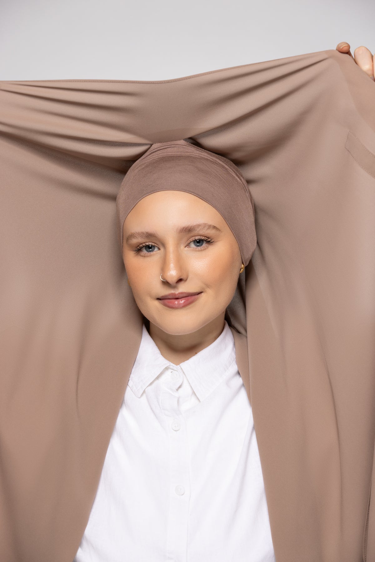 A woman wearing a taupe-beige headscarf and a white collared shirt, holding a matching taupe-beige fabric drape open above her head against a light gray studio background.